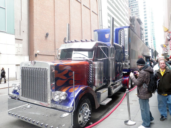 Optimus Prime and his trailer are parked on the street outside the showroom. Later, he'd get a parking ticket.